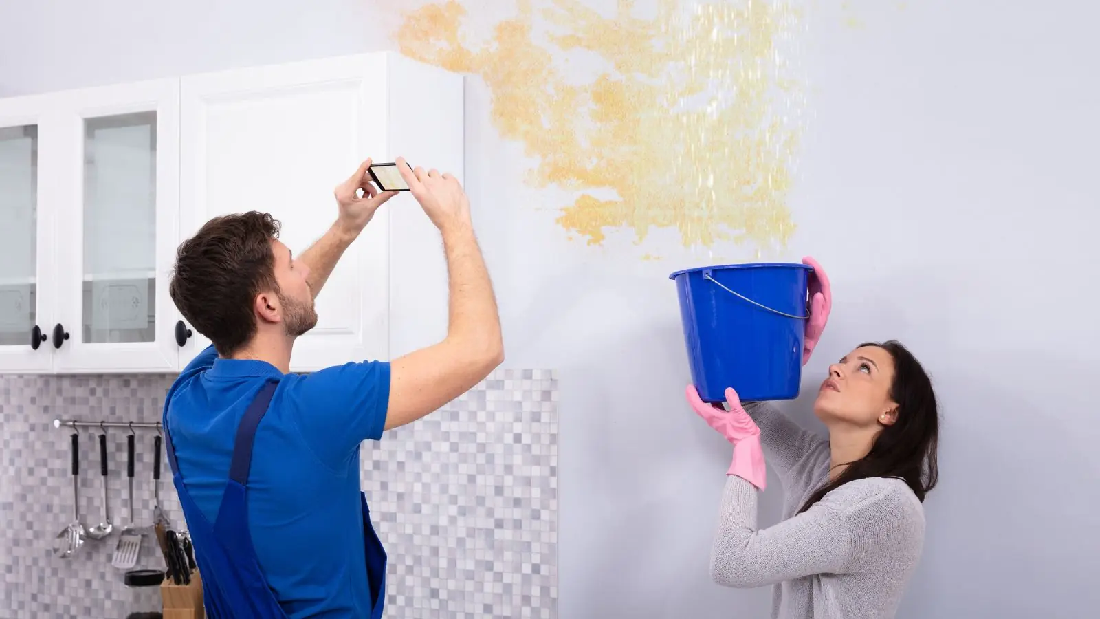 Homeowner photographing a ceiling leak while a second person holds a bucket